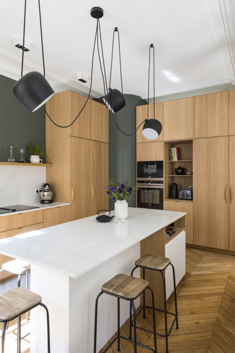 Une cuisine contemporaine dans un appartement haussmannien en bois avec un plan de travail et crédence blanc qui contraste avec un mur vert sapin